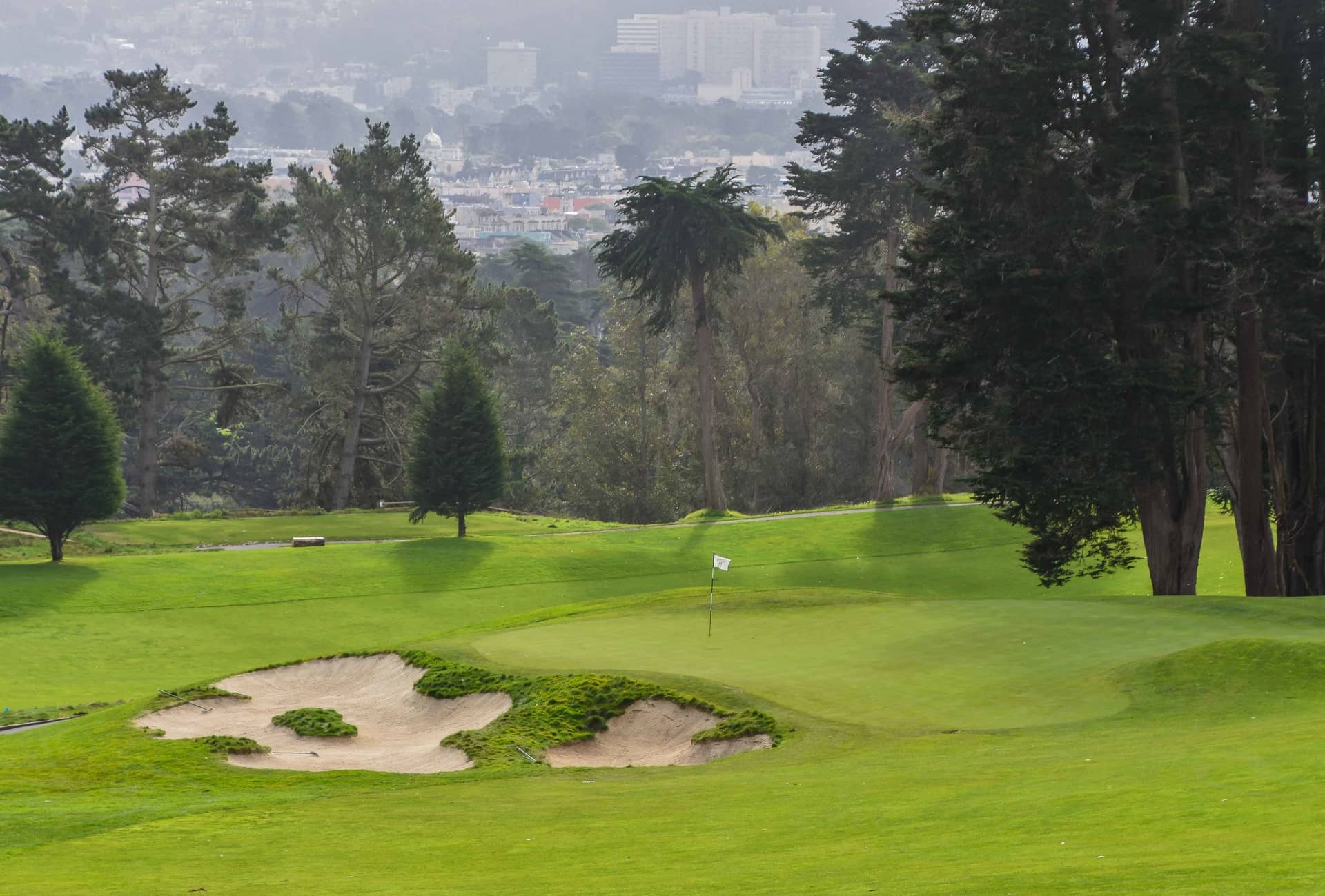 A Presidio Golf Course green bordered by bunkers, with San Francisco buildings beyond the trees. Photo by PJ Koenig / https://www.pjkoenig.com/presidio-golf-club